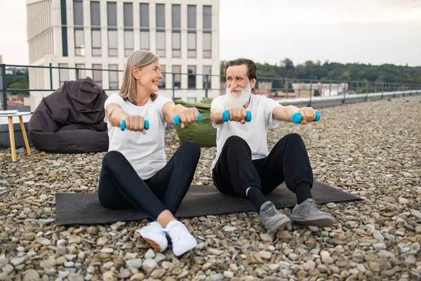 Yaşlı bir adam ve kadın çatıdaki minderlerde oturmuş halterlerle spor yapıyorlar..