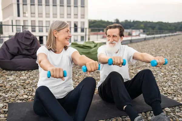 Üst düzey erkek ve kadın şehir merkezinde çatıda dambıllarla spor yapıyorlar.