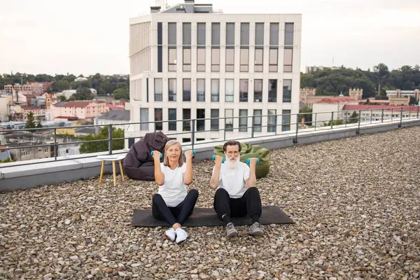 Üst düzey erkek ve kadın çatıdaki platformda oturuyorlar ve spor yapmak için halterleriyle egzersiz yapıyorlar.