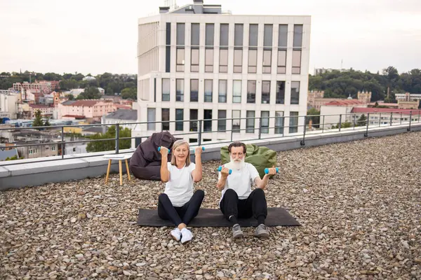 Yaşlı adam ve kadın, arka planda kentsel ufuk çizgisi olan dambıllarla spor yapıyor. Sağlıklı yaşam tarzı tanıtımı ve aktif emeklilik kavramı.