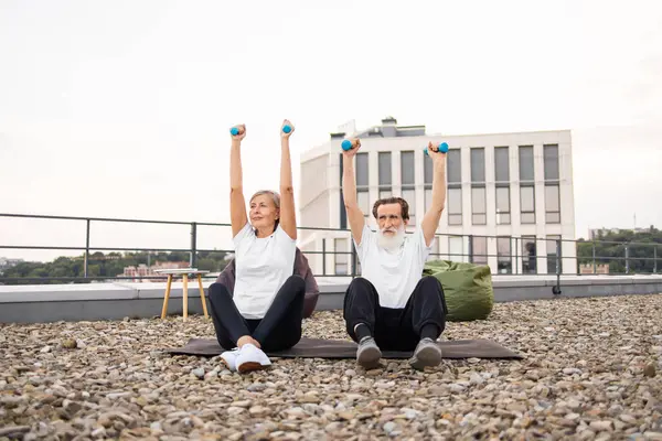 Yaşlı bir adam ve kadın spor yaparken dambılları kaldırıyor. Terasta oturuyorlar. Arka planda modern bir bina var.
