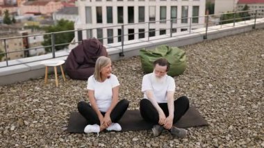Beyaz tişörtlü yaşlı adam ve kadın Lotus 'ta yoga meditasyonu yapıyorlar açık havada çatıda poz veriyorlar. Belirgin, sakin ve barışçıl ifadeler.