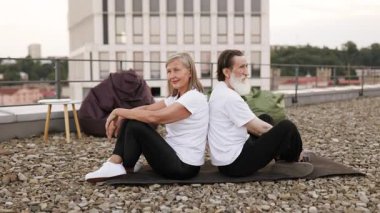 Üst düzey erkek ve kadın nilüfer çiçeğiyle yoga yapıyor çatıda poz veriyor rahatlama ve zindeliği birleştiriyor. Arka planda şehir manzarası görünüyor. Mankenler, sağlıklı yaşlanmayı temsil eden yaşlı erkek ve dişilerdir.