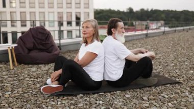 Yaşlı adam ve kadın sırt sırta oturuyorlar, meditasyon yogası, sakin bir atmosfer. Açık hava fitness, farkındalık egzersizi, sağlıklı yaşam tarzı, huzurlu akşam manzarası..