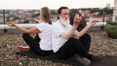 Üst düzey erkek ve kadın sırtüstü oturmuş çatıda Lotus pozunda meditasyon yapıyorlar. Farkındalık ve sağlık pratiğiyle meşgul olmak. Yaşlı yetişkinler için sağlıklı yaşam biçimi, dinlenmek için huzurlu şehir ortamı.