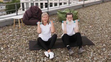 Üst düzey çift dambıllarıyla çatıda egzersiz yapıyor. Yaşlı adam ve kadın dışarıda spor yapıyorlar..