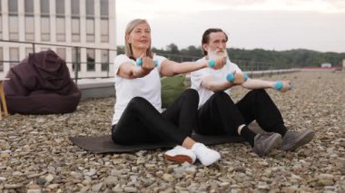 Yaşlı bir adam ve kadın çatıdaki minderlerde oturmuş halterlerle spor yapıyorlar..
