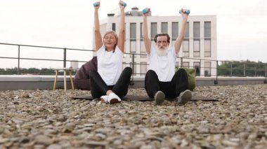 Yaşlı bir adam ve kadın spor yaparken dambılları kaldırıyor. Terasta oturuyorlar. Arka planda modern bir bina var.