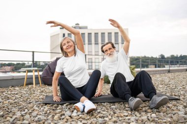 Yaşlı bir adam ve kadın birlikte egzersiz yapıyor, dışarıda yoga yaparken kollarını esnetiyorlar. Sağlıklı yaşam tarzı, canlılık, açık hava egzersizi terasta olgun yetişkinler için esneklik ve gevşemeyi teşvik ediyor.