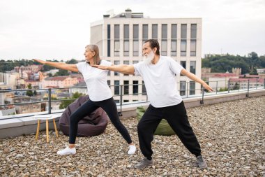 Üst düzey erkek ve kadın çatıda yoga yapıyor, şehir ortamında savaşçı pozu veriyorlar. Sağlıklı yaşam tarzı, açık hava fitness aktivitesi fiziksel sağlık ve zihinsel refahı teşvik, enerji gösterisi