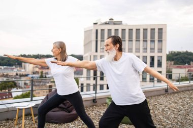 Üst düzey erkek ve kadın, şehir manzarasına tepeden bakan çatı katında yoga yapıyorlar. İkisi de gündelik giysiler ve rahat giysiler giyip savaşçı yoga pozları veriyor ve dengeyi vurguluyor..