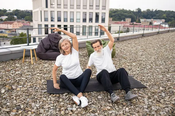 Üst düzey erkek ve kadın çatıda paspaslar üzerinde esnetiyorlar, fitness aktivitesine katılıyorlar, esnekliği ve sağlığı vurguluyorlar..