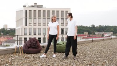 Üst düzey erkek ve kadın çatıda yoga yapıyor, şehir ortamında savaşçı pozu veriyorlar. Sağlıklı yaşam tarzı, açık hava fitness aktivitesi fiziksel sağlık ve zihinsel refahı teşvik, enerji gösterisi