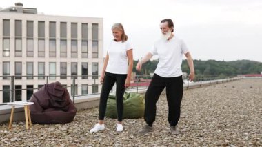 Üst düzey erkek ve kadın, şehir manzarasına tepeden bakan çatı katında yoga yapıyorlar. İkisi de gündelik giysiler ve rahat giysiler giyip savaşçı yoga pozları veriyor ve dengeyi vurguluyor..