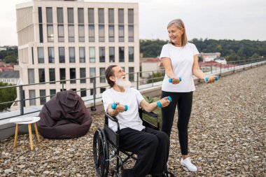 Yaşlı kadın, tekerlekli sandalyedeki yaşlı adama dışarıda spor yaparken yardım ediyor mavi halterlerle iyileşme ve refahı arttırıyor..