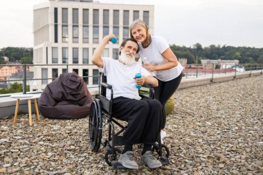 Tekerlekli sandalyedeki yaşlı adam dışarıda halterlerle egzersiz yapıyor, gülümseyen olgun bir kadın tarafından destekleniyor.
