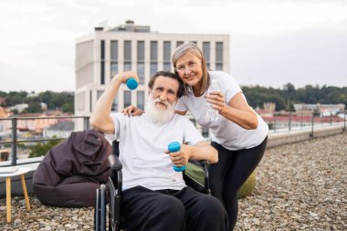 Son sınıf çifti terasta spor yapıyor. Dambılı yaşlı bir adam su tutan bir kadın tarafından destekleniyor, sağlık, arkadaşlık, aktif yaşam tarzı ve olgun bireyler için rehabilitasyon destekliyor..