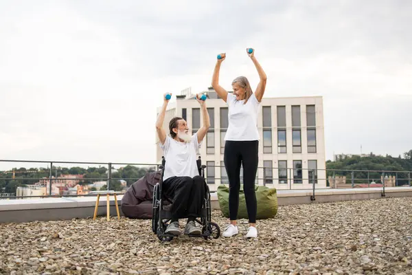 Tekerlekli sandalyedeki son sınıf öğrencisi ve şehir çatılarında dambıllarıyla egzersiz yapan aktif yaşam tarzlarını destekleyen kadın. Cesaret, birliktelik, sağlık, canlılık ve fitness desteği