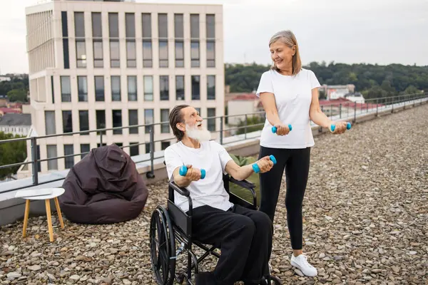Yaşlı kadın, tekerlekli sandalyedeki yaşlı adama dışarıda spor yaparken yardım ediyor mavi halterlerle iyileşme ve refahı arttırıyor..