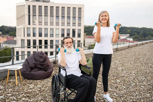 Yaşlı bir çift halterlerle hafif egzersizler yapıyor, iyileşme ve sağlığa teşvik ediyorlar. Kadın, tekerlekli sandalyede oturan kocasına yardım ederken bir şeyler gösteriyor. Fitness ve rehabilitasyonu vurguluyor