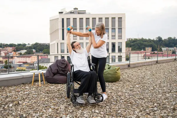 Yaşlı kadın tekerlekli sandalyeye bağlı kocasına rehabilitasyon egzersizleri için çatıdaki dambılları kullanarak yardım ediyor. Parlak gün, destekleyici, duygusal etkileşim, iyileşmeyi vurgulamak, birliktelik.