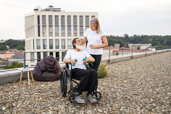 Yaşlı çift dışarıda birlikte egzersiz yapıyor. Kocasını destekleyen yaşlı kadın fiziksel aktivite sırasında tekerlekli sandalyede oturmuş iyileşme ve güç odaklı..