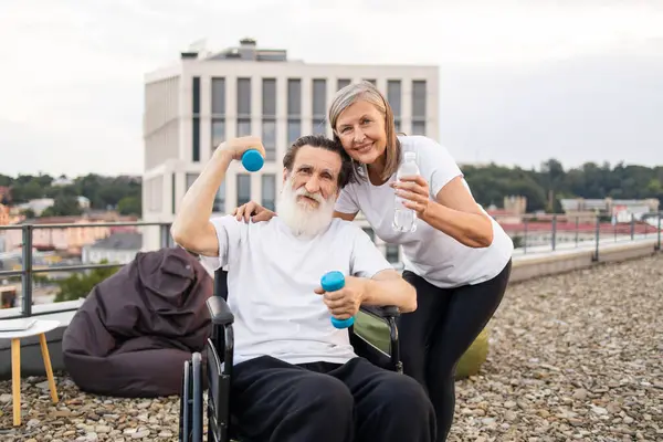 Son sınıf çifti terasta spor yapıyor. Dambılı yaşlı bir adam su tutan bir kadın tarafından destekleniyor, sağlık, arkadaşlık, aktif yaşam tarzı ve olgun bireyler için rehabilitasyon destekliyor..