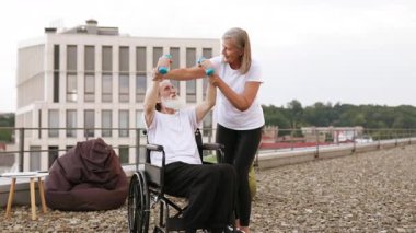 Yaşlı bir kadın yaşlı bir kocaya yardım ederken tekerlekli sandalye bağlı, açık hava gösterileri sunuyor fiziksel kısıtlamalara rağmen karşılıklı destek ve sağlıklı yaşam tarzı tanıtımı yapıyor..