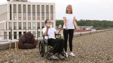 Tekerlekli sandalyedeki yaşlı adam çatıdaki kadının yardımıyla dambıllarla egzersiz yapıyor. Açık havada spor yapmayı seviyorlar, kapsayıcı çevredeki sağlık ve rehabilitasyona odaklanıyorlar..