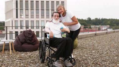 Son sınıf çifti terasta spor yapıyor. Dambılı yaşlı bir adam su tutan bir kadın tarafından destekleniyor, sağlık, arkadaşlık, aktif yaşam tarzı ve olgun bireyler için rehabilitasyon destekliyor..