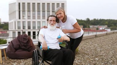 Tekerlekli sandalyedeki yaşlı adam dışarıda halterlerle egzersiz yapıyor, gülümseyen olgun bir kadın tarafından destekleniyor.