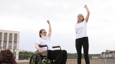 Tekerlekli sandalyede yaşlı bir adam ve kadın yan yana durmuş şehir çatısı dışında kollarını kaldırıyor.