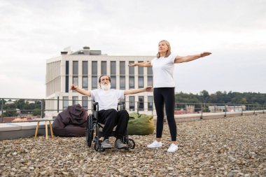 Yaşlı bir adam yaşlı bir kadının yanında tekerlekli sandalye kullanıyor. Çatıda kollarını açıyor..