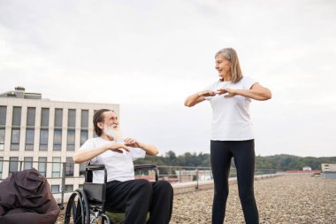 Yaşlı bir kadın, tekerlekli sandalyedeki adama çatıda egzersiz yaptırıyor, spor ve refahı teşvik ediyor. Kadın ve erkek takım çalışması, kapsayıcılık ve aktif yaşam tarzı gösteriyor..