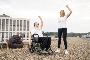 Tekerlekli sandalyedeki yaşlı adam ve kadın dışarıda esneme egzersizleri yapıyor, birlikte aktiviteleri teşvik ediyorlar..