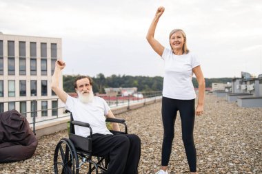 Tekerlekli sandalyede yaşlı bir adam ve kadın yan yana durmuş şehir çatısı dışında kollarını kaldırıyor.