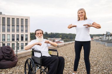 Tekerlekli sandalyede engelli yaşlı bir adam ve çatıda kollarını açan yaşlı bir kadın. Yaşlı yetişkinler için aktif yaşam tarzını destekleyen sağlık ve sağlık faaliyetleri.