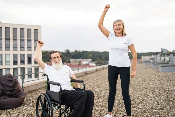 Tekerlekli sandalyede yaşlı bir adam ve kadın yan yana durmuş şehir çatısı dışında kollarını kaldırıyor.