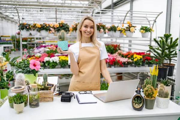 Beyaz bir kadın çiçekçi çiçeklerle dolu bir dükkanda kaktüs tutarken gülümsüyor..