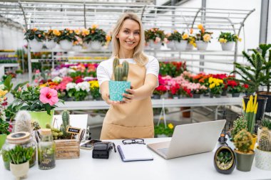 Çiçekçi dükkanında beyaz bir kadın elinde kaktüs, bitkilerle dolu bir satış masasının arkasında kameraya gülümsüyor..