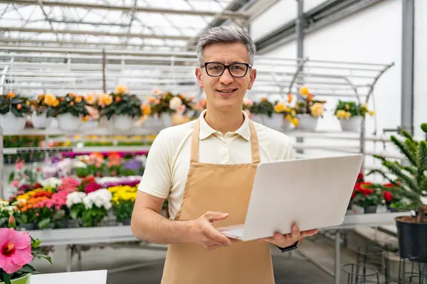 Elinde dizüstü bilgisayar olan bir çiçekçi, canlı çiçekçi dükkanından gülümsüyor, hoş bir ortam sunuyor..