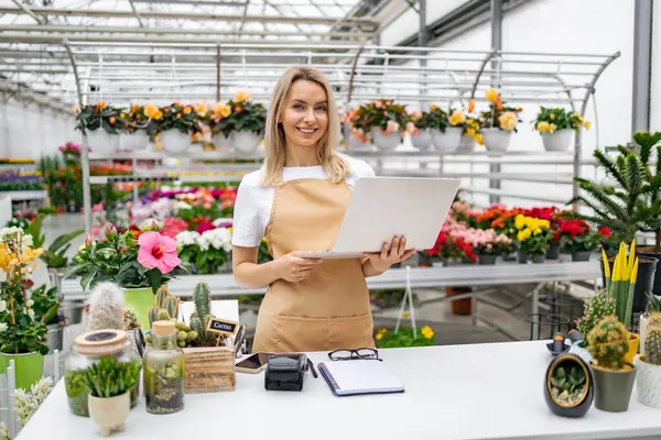 Dükkanda dizüstü bilgisayarı olan beyaz bir kadın çiçekçi. Etrafı bitkilerle çevrili, içerikli ve müşterilere yardım etmeye hazır..