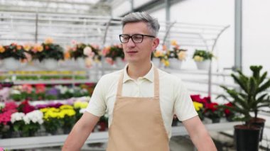 Çiçekçisinde kollarını kavuşturmuş beyaz bir adam. Satılık çiçek ve saksı bitkileriyle çevrili..