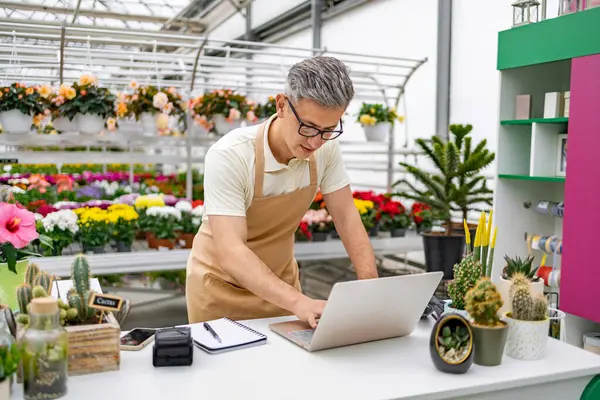 Çiçekçi dükkanındaki beyaz bir adam, bitki ve çiçeklerle çevrili bir tezgahta dizüstü bilgisayar kullanıyor. Envanter ya da satış işlerini yönetiyor gibi görünüyor..