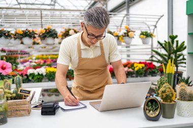 Beyaz bir adam, bir çiçekçi, dizüstü bilgisayarda çalışır ve arka planda çeşitli bitkiler ve çiçeklerle mağaza masasında notlar alır..