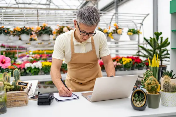 Beyaz bir adam, bir çiçekçi, dizüstü bilgisayarda çalışır ve arka planda çeşitli bitkiler ve çiçeklerle mağaza masasında notlar alır..