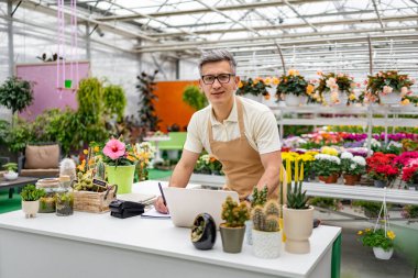 Beyaz bir adam çiçekçi tezgahında çalışıyor. Etrafı canlı bitki ve çiçeklerle çevrili..
