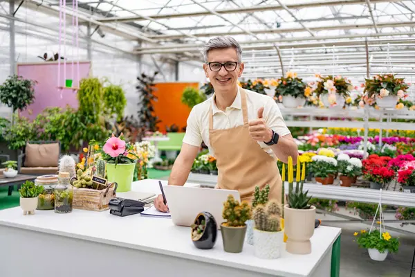 Beyaz bir adam çiçekçi tezgahında çalışıyor. Etrafı canlı bitki ve çiçeklerle çevrili..