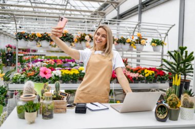 Beyaz bir kadın çiçekçi dükkanında selfie çekiyor. Etrafı canlı çiçekler ve bitkilerle çevrili..