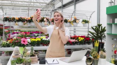Gülümseyen beyaz bir kadın, canlı bitkilerle çevrili parlak, iyi stoklanmış çiçekçisinde selfie çekiyor..